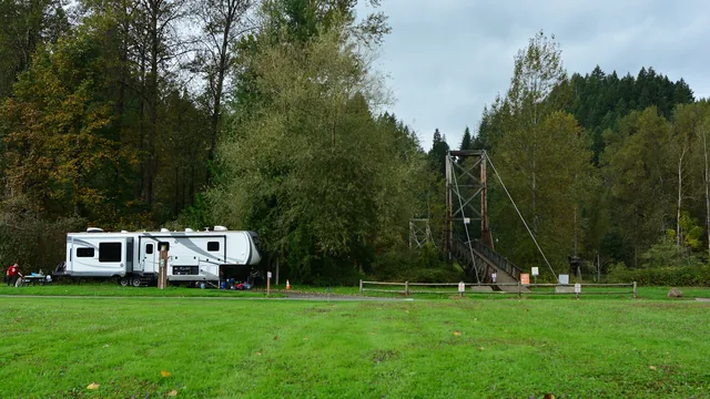 A long fifth-wheel trailer set up at the edge of a mown field at Tolt-MacDonald Park in Washington, a suspension footbridge crossing the river behind it.